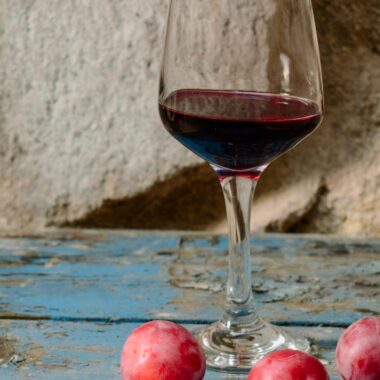 A glass of red wine alongside fresh plums on a rustic wooden table.