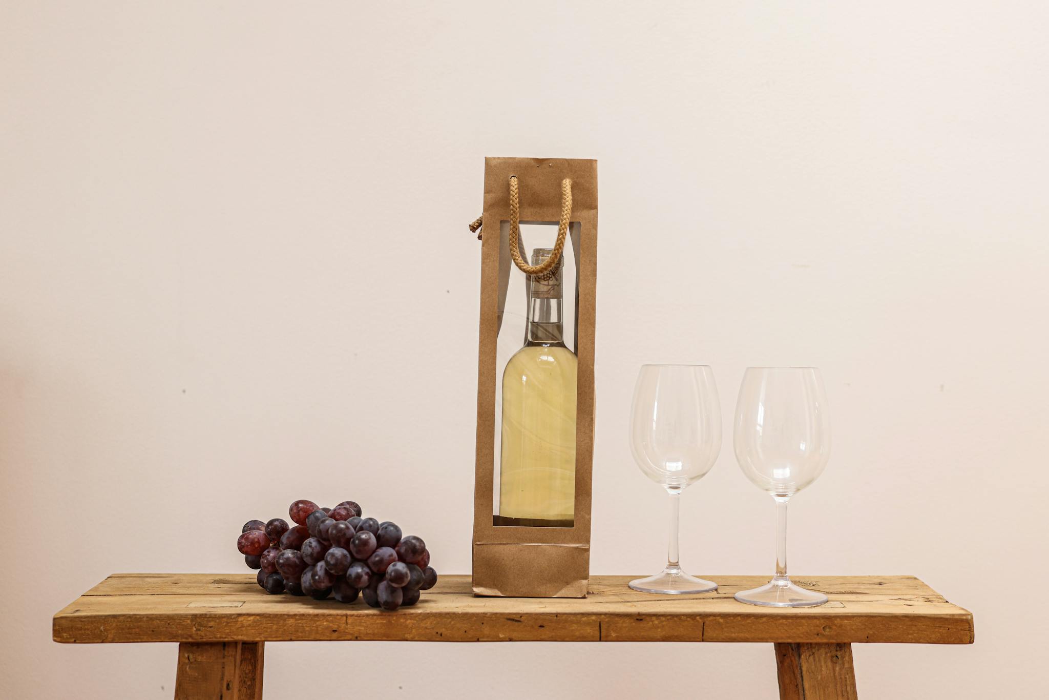 A sophisticated setup featuring a bottle of white wine in a paper bag, grapes, and wine glasses on a wooden table.